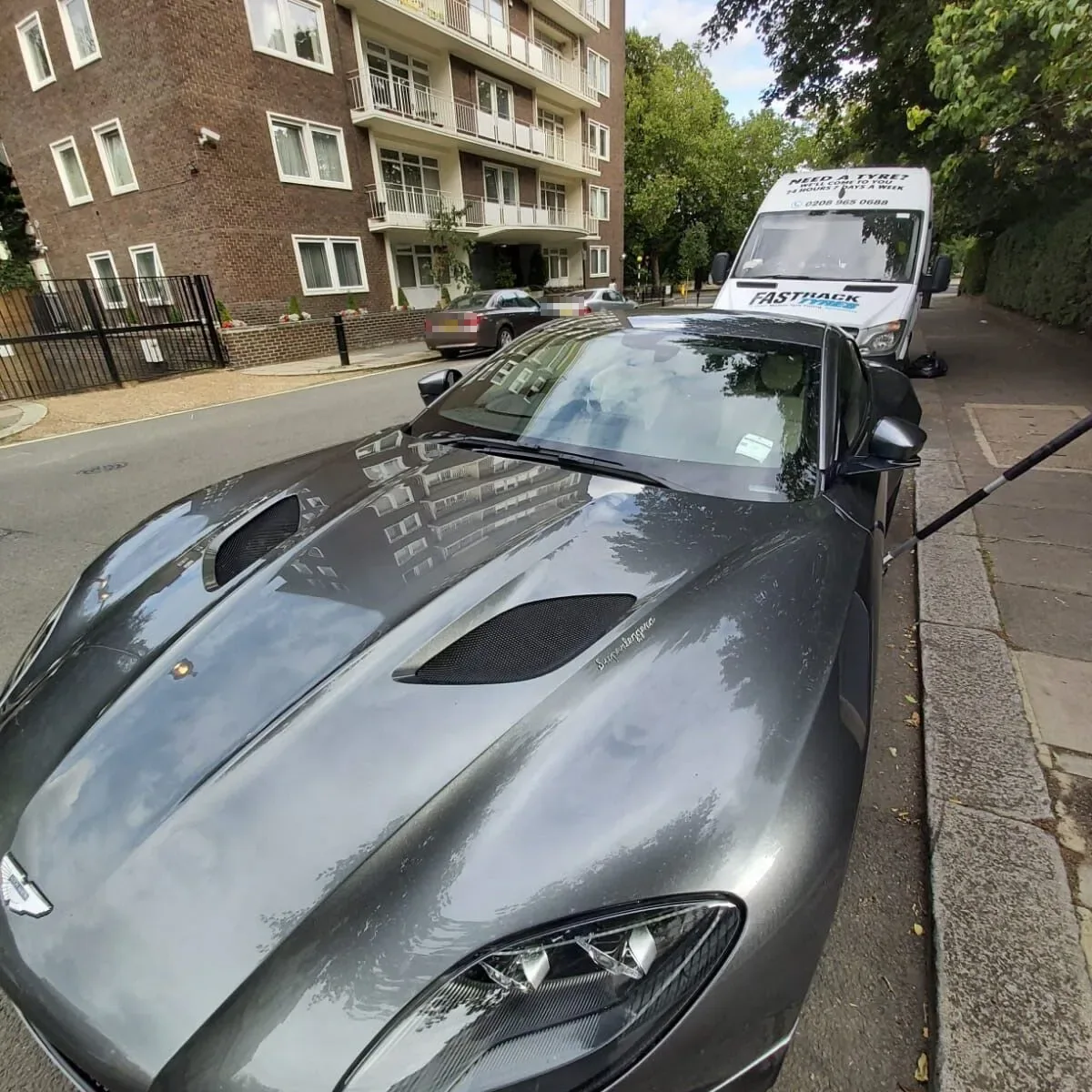 Grey Aston Martin Superleggera with its bonnet to camera and the Fastrack Tyres van behind on a London street