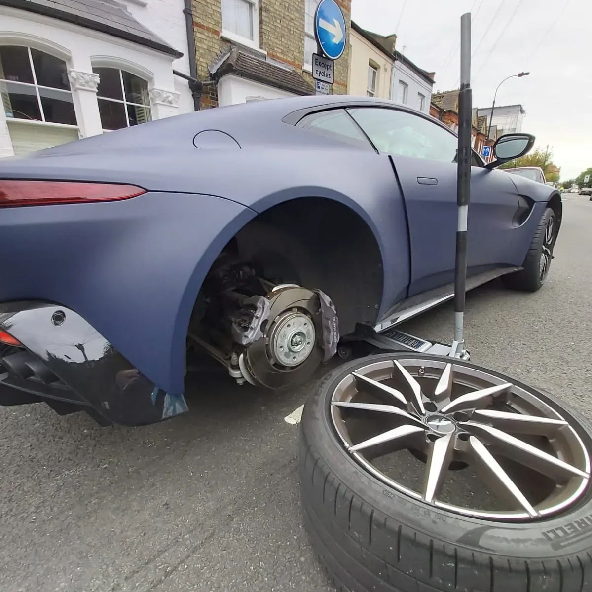 Matte navy-blue Aston Martin Vantage with its rear wheel removed and the alloy resting on the road during a roadside tyre change