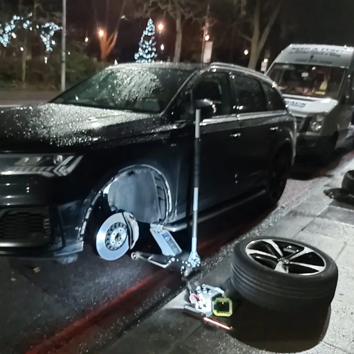 Black Audi Q7 jacked up at night beside a lit Christmas tree with the front wheel removed and the spare alloy on the pavement