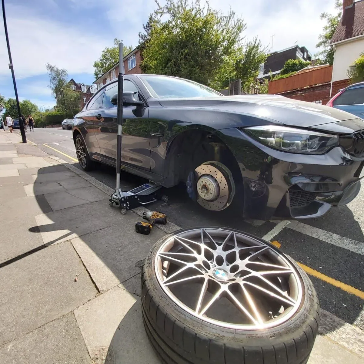 Black BMW M4 on a jack with its star-spoke M alloy wheel resting in the foreground during a roadside tyre change