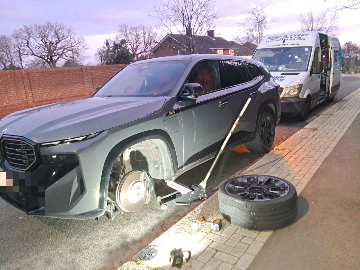 Grey BMW XM performance SUV with a wheel off at dusk for a mobile tyre replacement, Fastrack van behind