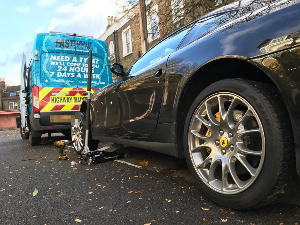 Black Ferrari 612 Scaglietti raised on a trolley jack for a wheel change with the Fastrack Tyres van behind