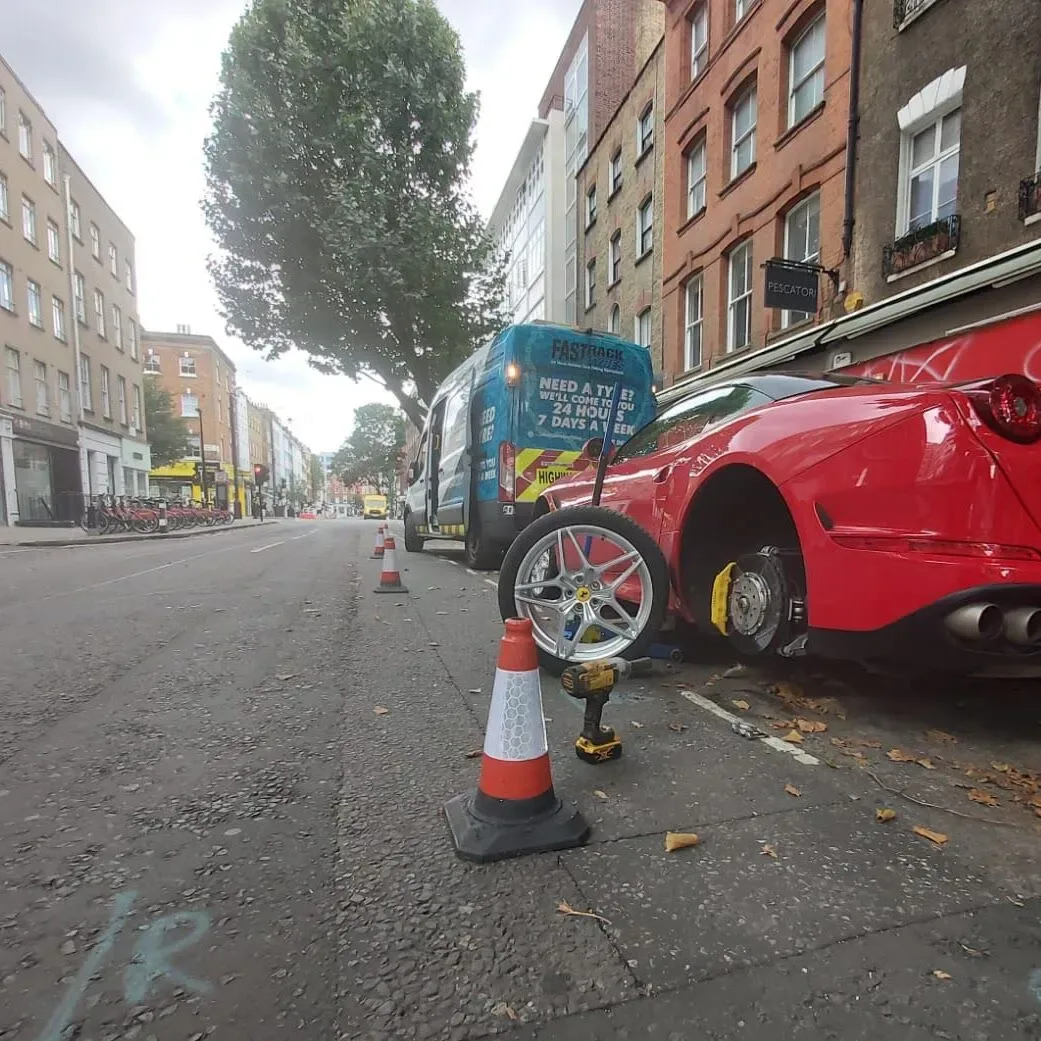 Red Ferrari having a rear wheel changed on a London street with traffic cones out and the Fastrack Tyres van behind