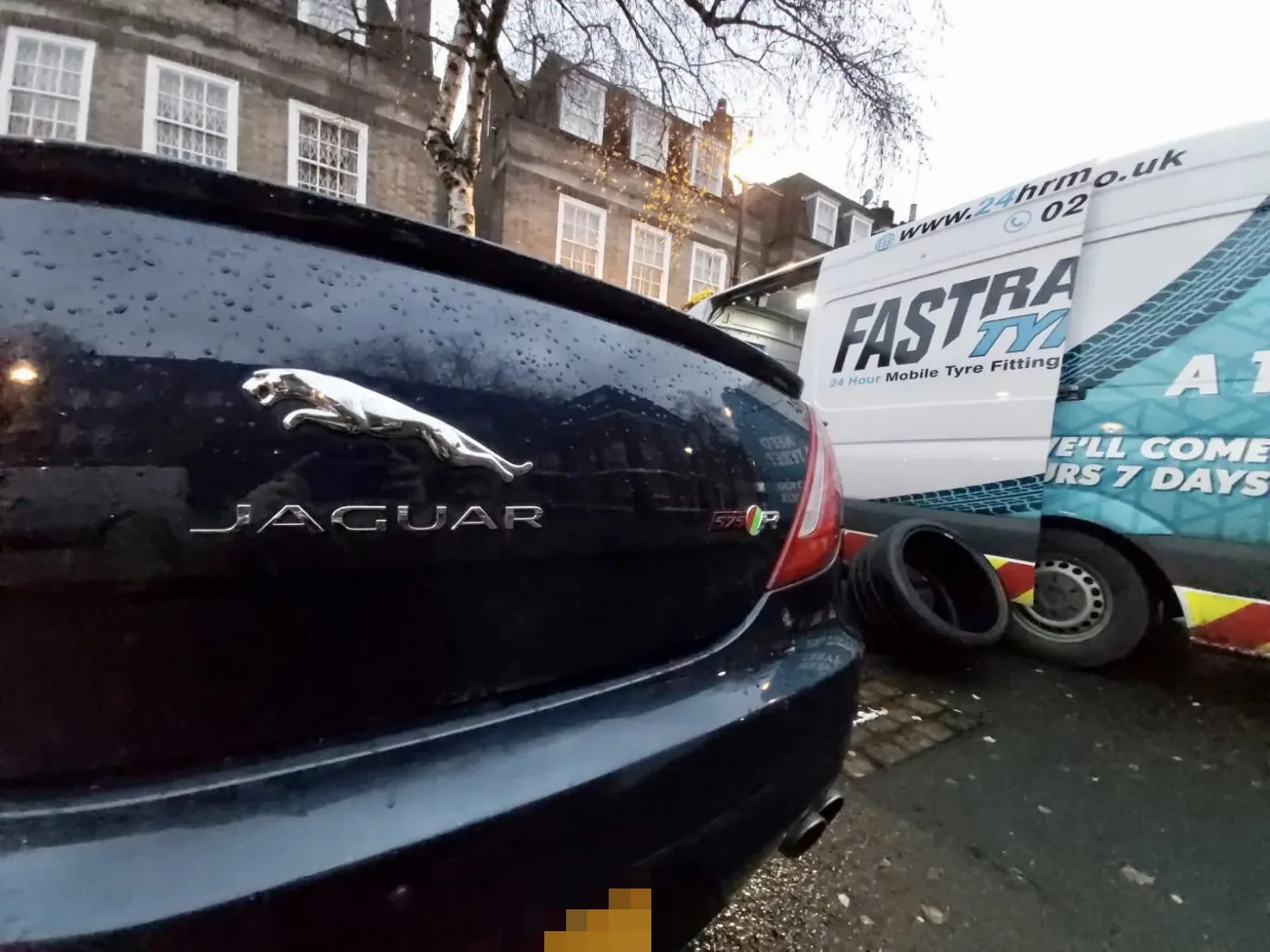 Black Jaguar XJR 575 on a wet London street at dusk with the Fastrack Tyres van behind during a wheel change