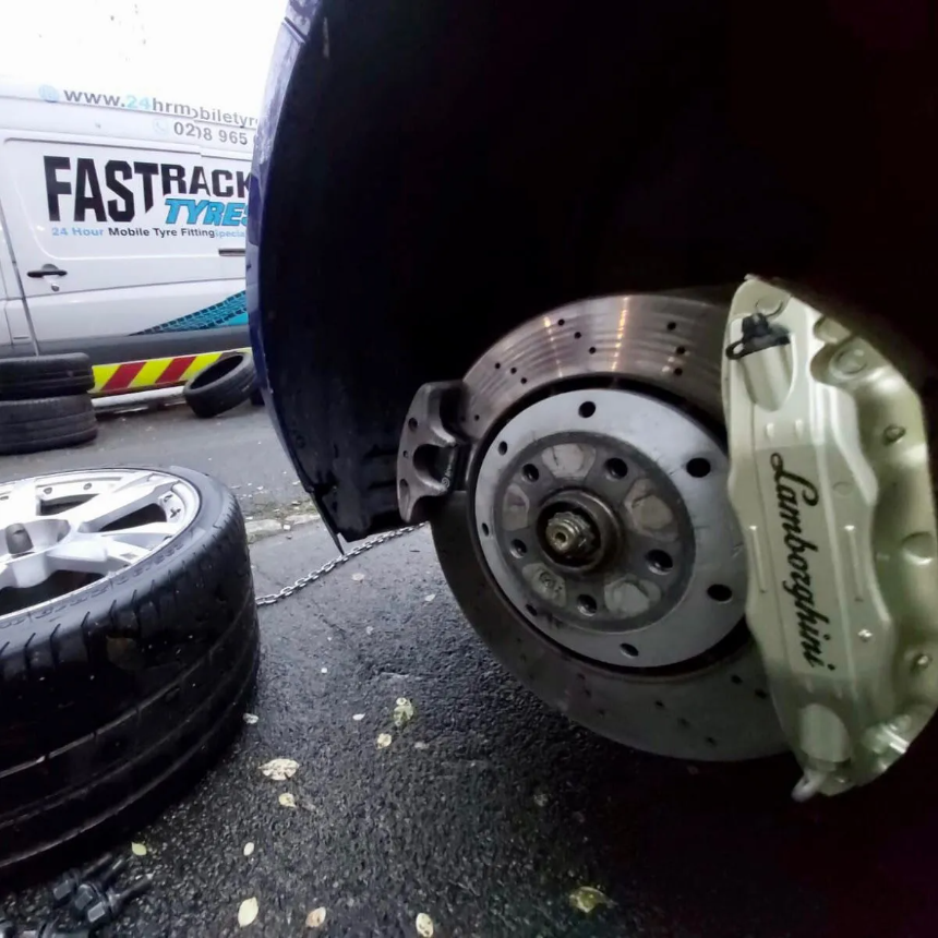 Close-up of a Lamborghini cross-drilled brake disc and cream caliper with the wheel off and the Fastrack van behind