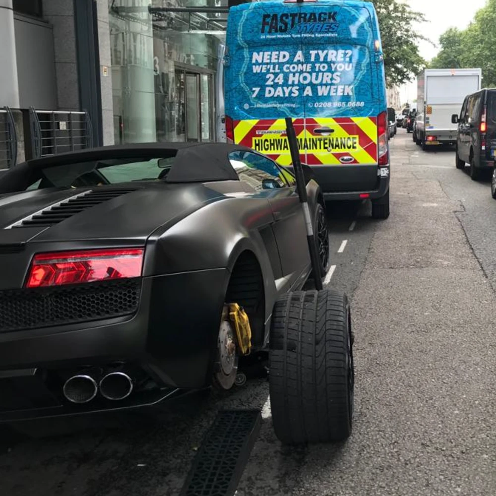 Matte-black Lamborghini Gallardo Spyder with its rear wheel removed for a mobile tyre change on a Mayfair street, the branded Fastrack Tyres van parked behind