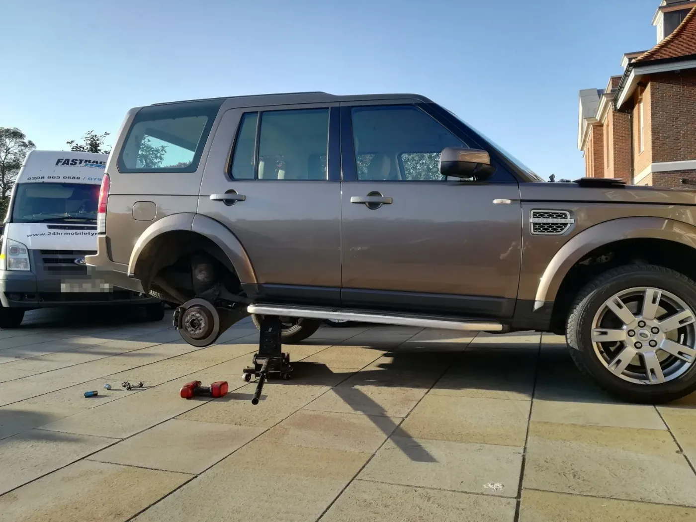 Bronze Land Rover Discovery 4 raised on a trolley jack with its wheel removed on a driveway, the Fastrack Tyres van behind