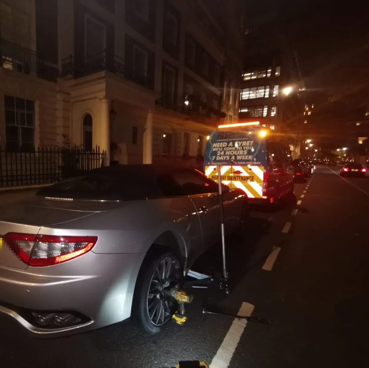 Silver Maserati GranTurismo convertible with its rear wheel removed at night in Belgravia, the Fastrack Tyres van behind