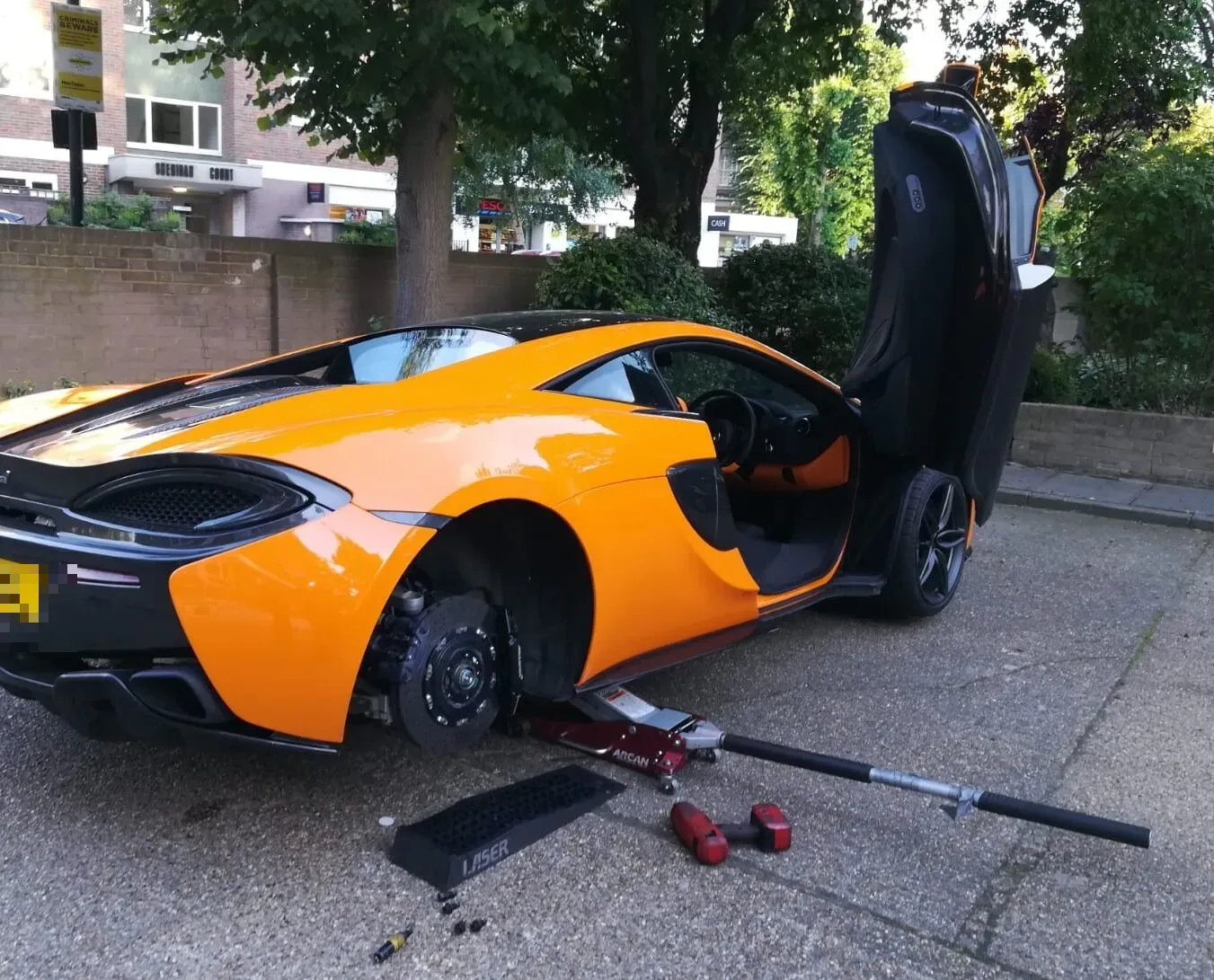 Orange McLaren 570S with its dihedral door raised having a wheel changed on a London street
