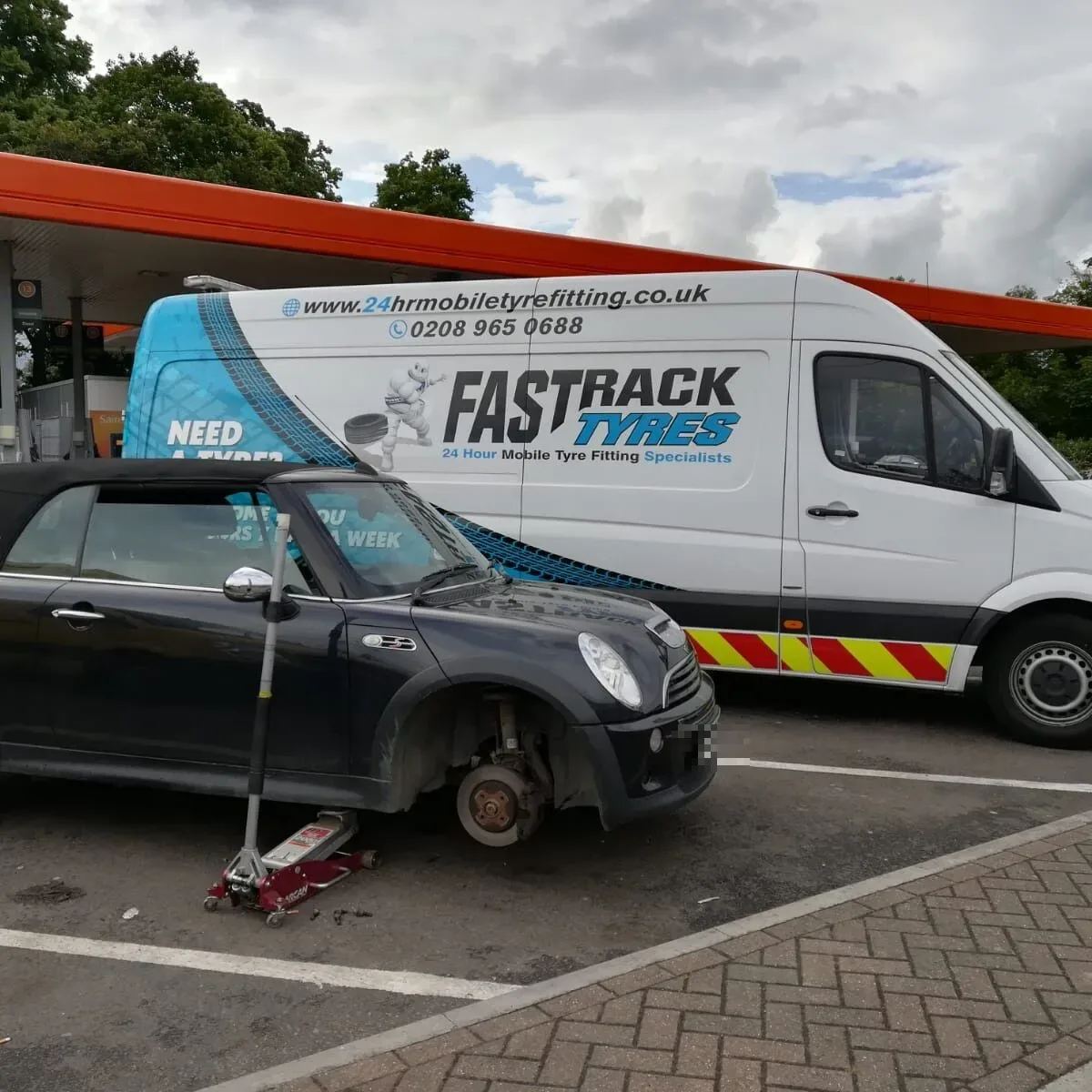 Black Mini convertible with its wheel removed on a petrol station forecourt with the Fastrack Tyres van behind