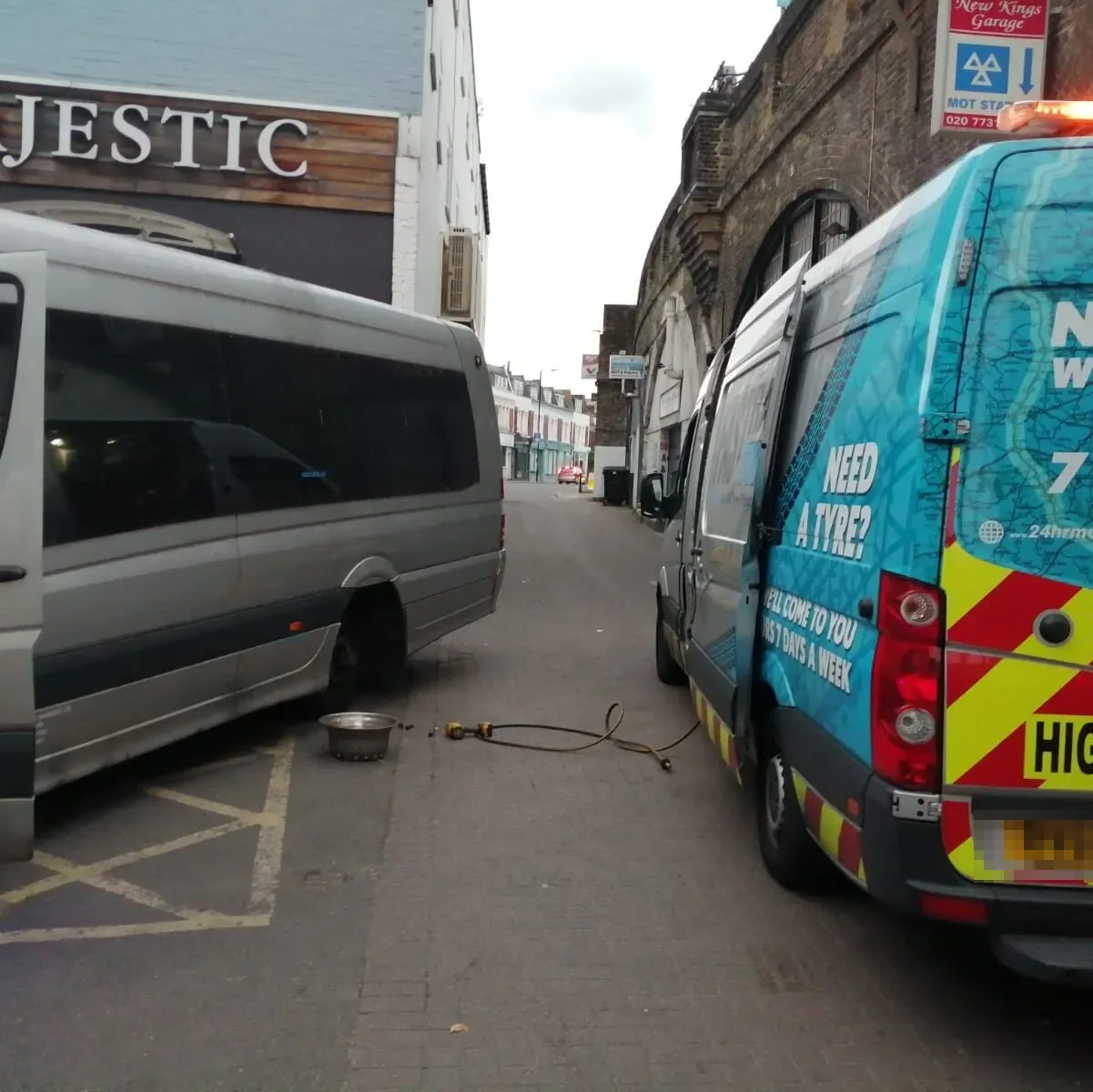 Silver minibus with a flat rear tyre being worked on beside the Fastrack Tyres van on a London side street