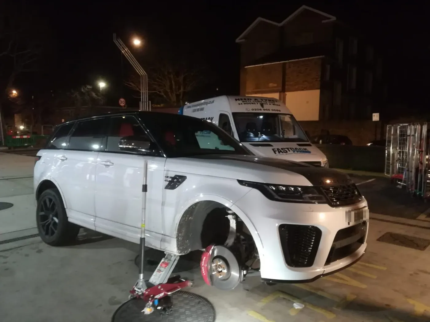 White Range Rover Sport SVR raised on a trolley jack with its front wheel removed at night, the Fastrack Tyres van behind