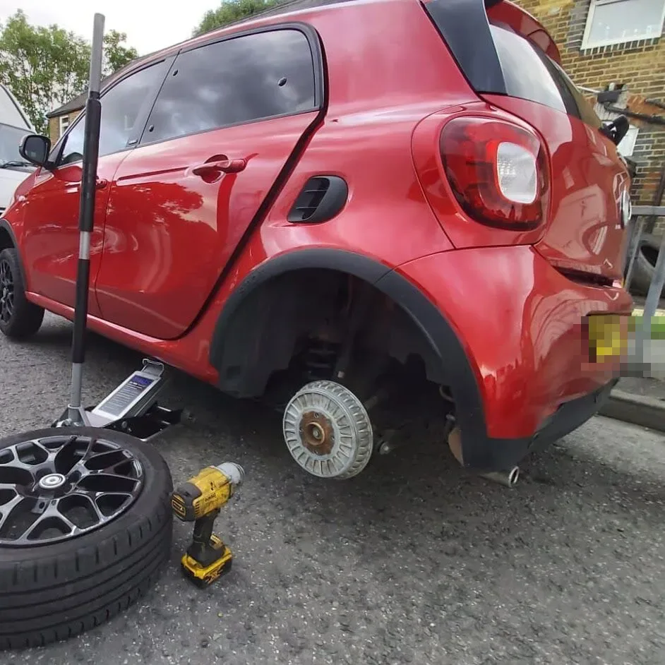 Red Smart ForFour raised on a trolley jack with its rear wheel removed and the alloy resting on the road during a roadside tyre change