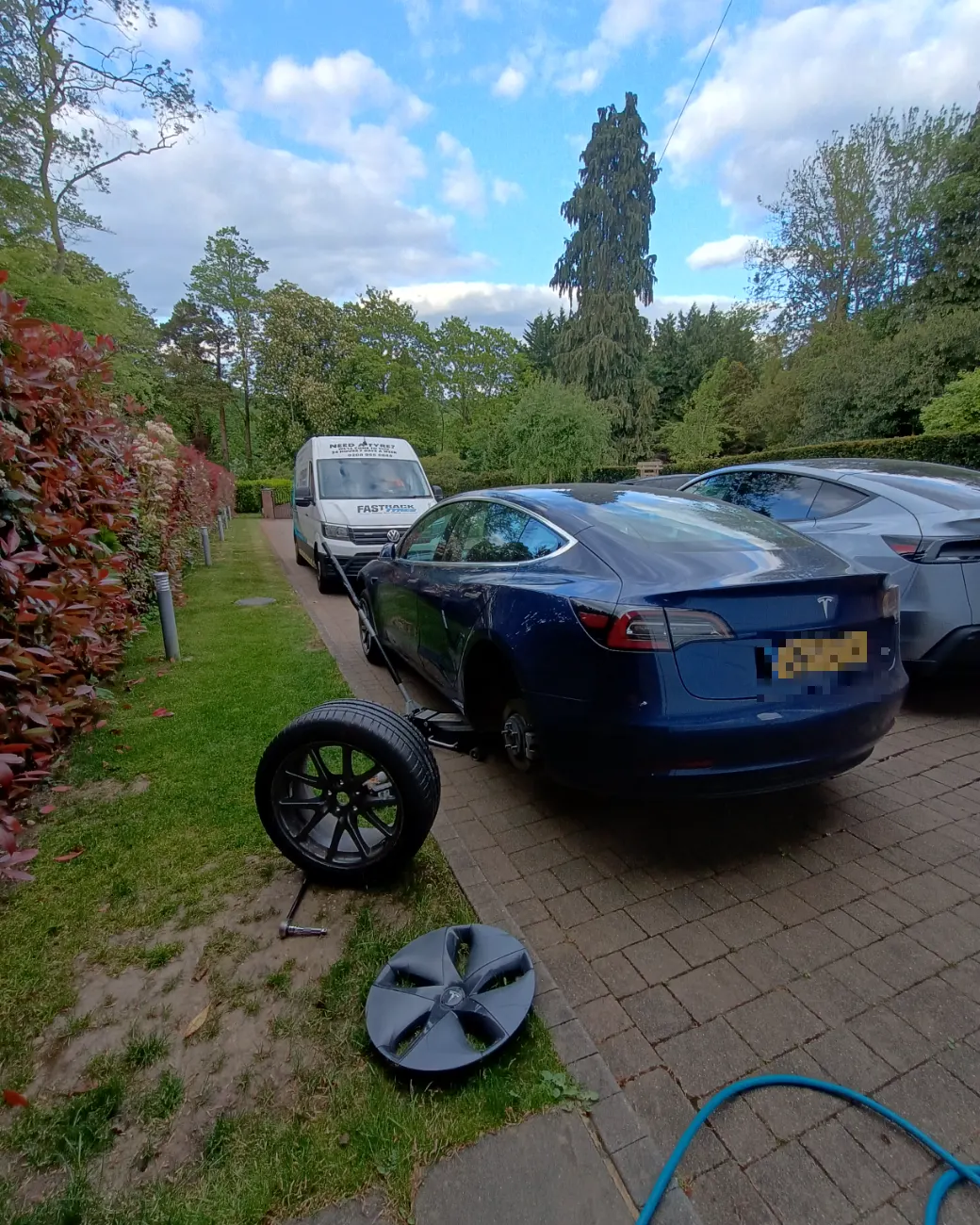 Blue Tesla Model 3 having a wheel changed on a leafy private driveway